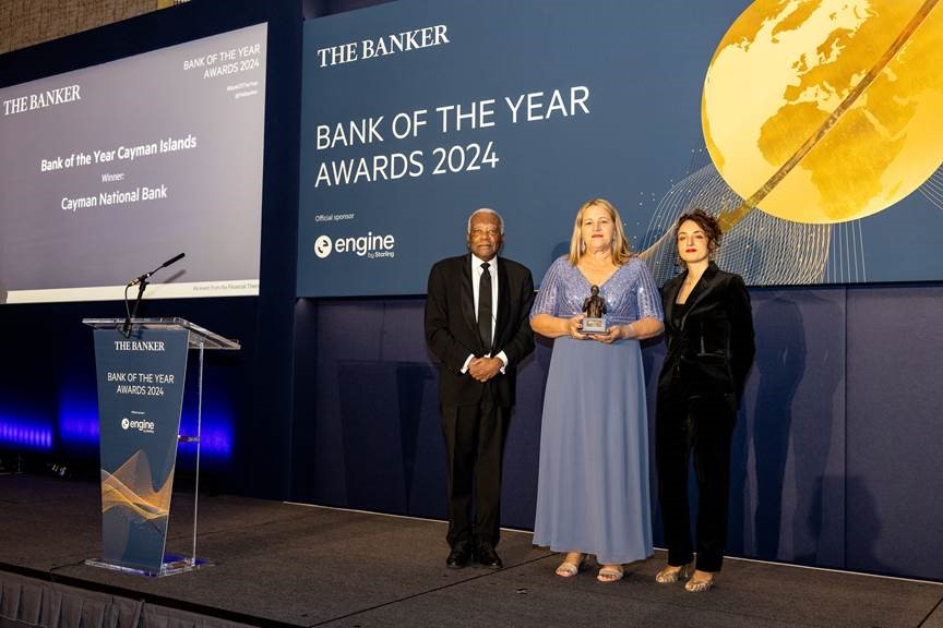 Three people stand on stage at the Bank of the Year Awards 2024, holding a trophy. A banner reads "Bank of the Year Cayman Islands - Cayman National Bank.