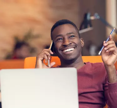 Man smiling while talking on his smartphone and sitting at his desk with his laptop